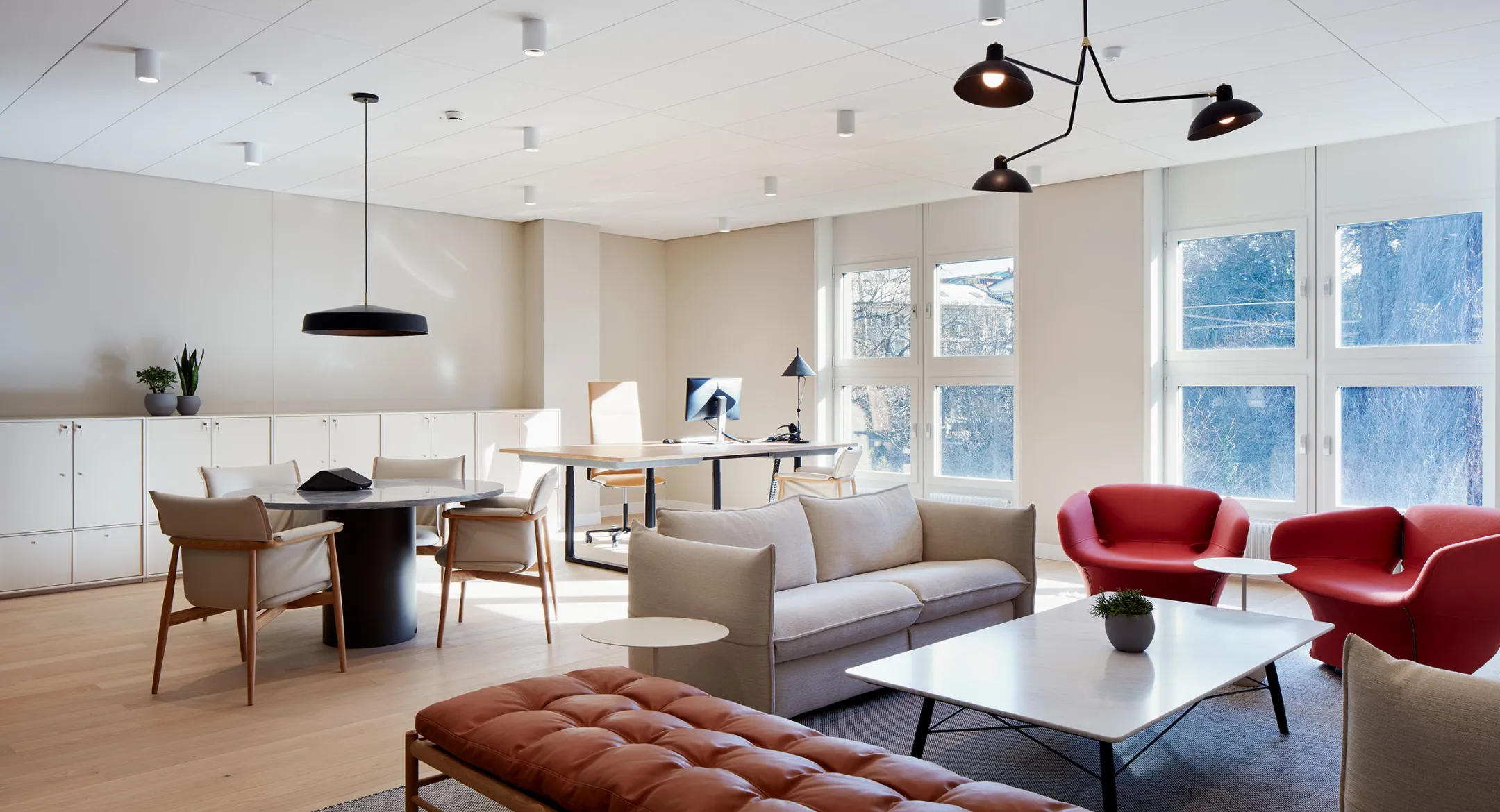 Bright modern office lounge with beige sofa, two red armchairs, round and rectangular white tables, and a workspace with a desk and computer by large windows.
