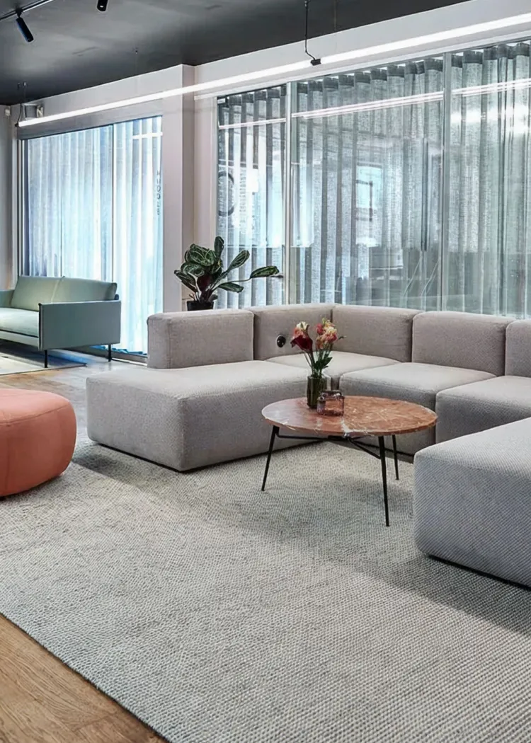 Modern office lounge with light gray sectional sofa, round marble coffee table with flowers, and green armchair near large glass windows with vertical blinds.