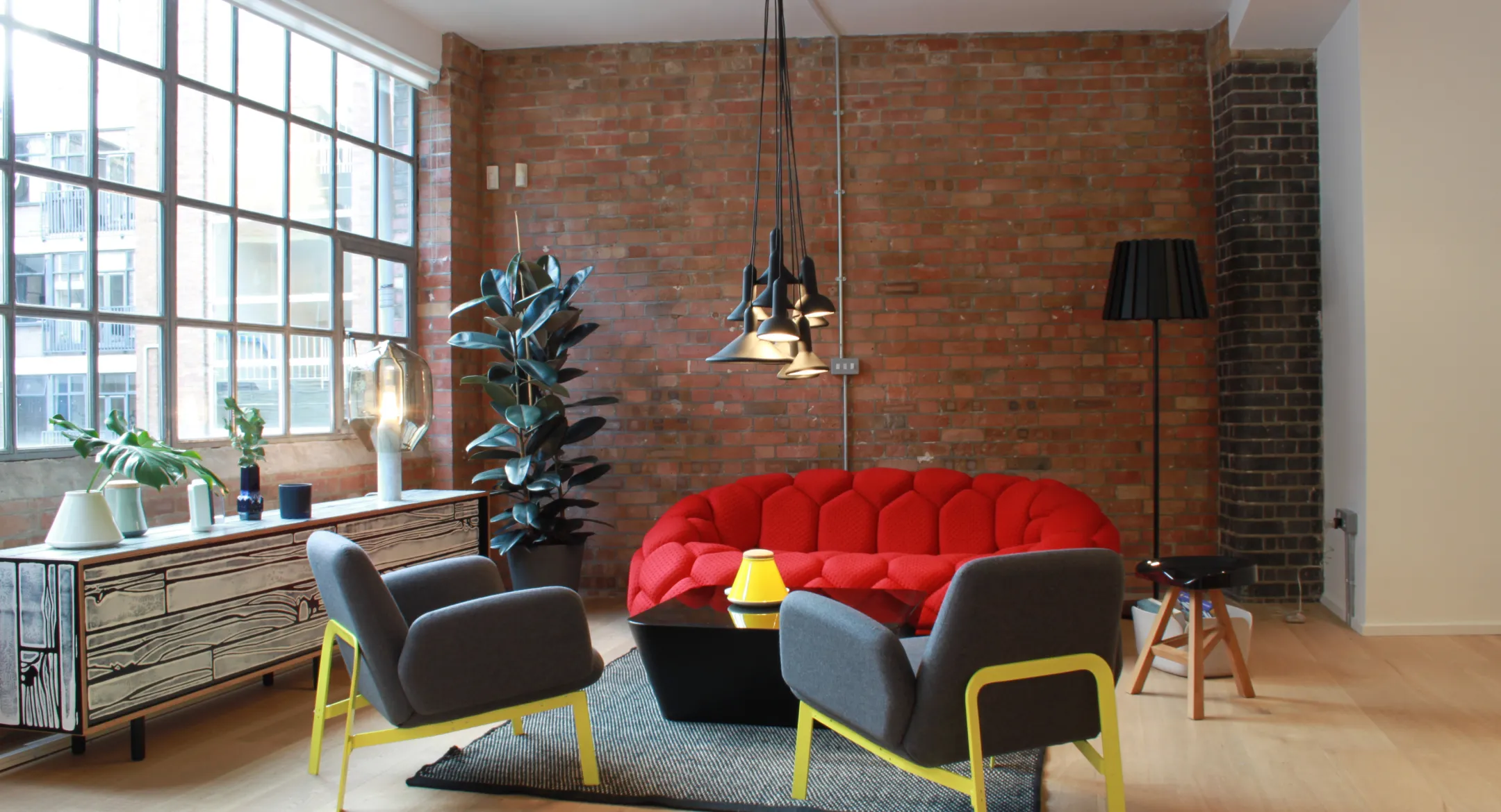 Modern living room with a red tufted sofa, two gray chairs with yellow legs, a black coffee table, large window, and brick wall.