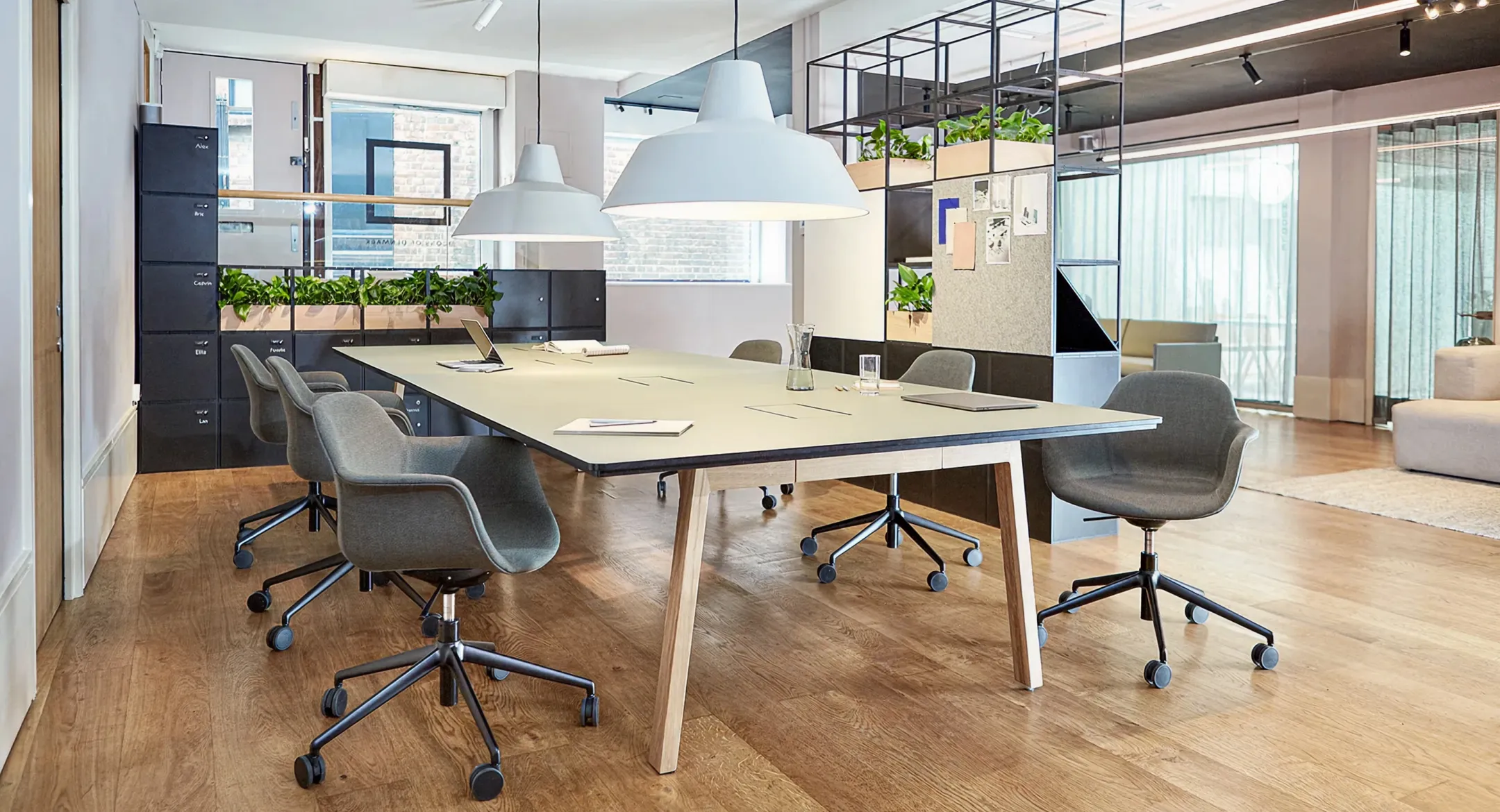Modern conference room with a rectangular table surrounded by gray swivel chairs, large pendant lights, and green plants on shelves.