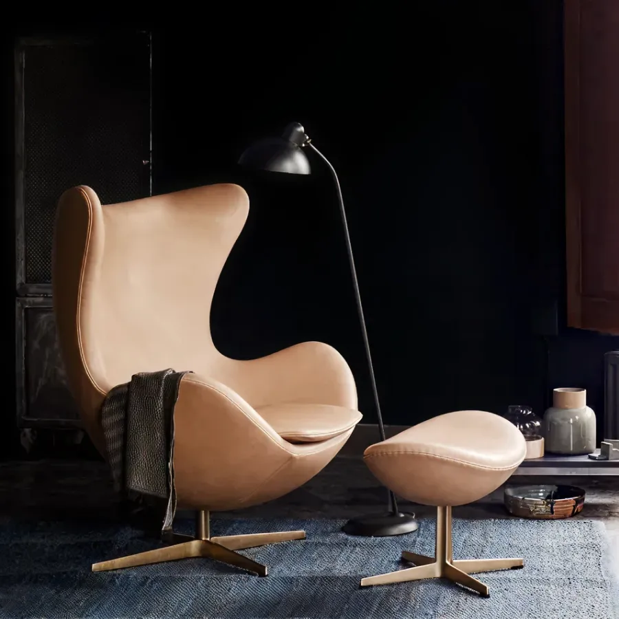 Modern tan leather lounge chair with matching ottoman on a blue rug, next to a black floor lamp and dark wall background.
