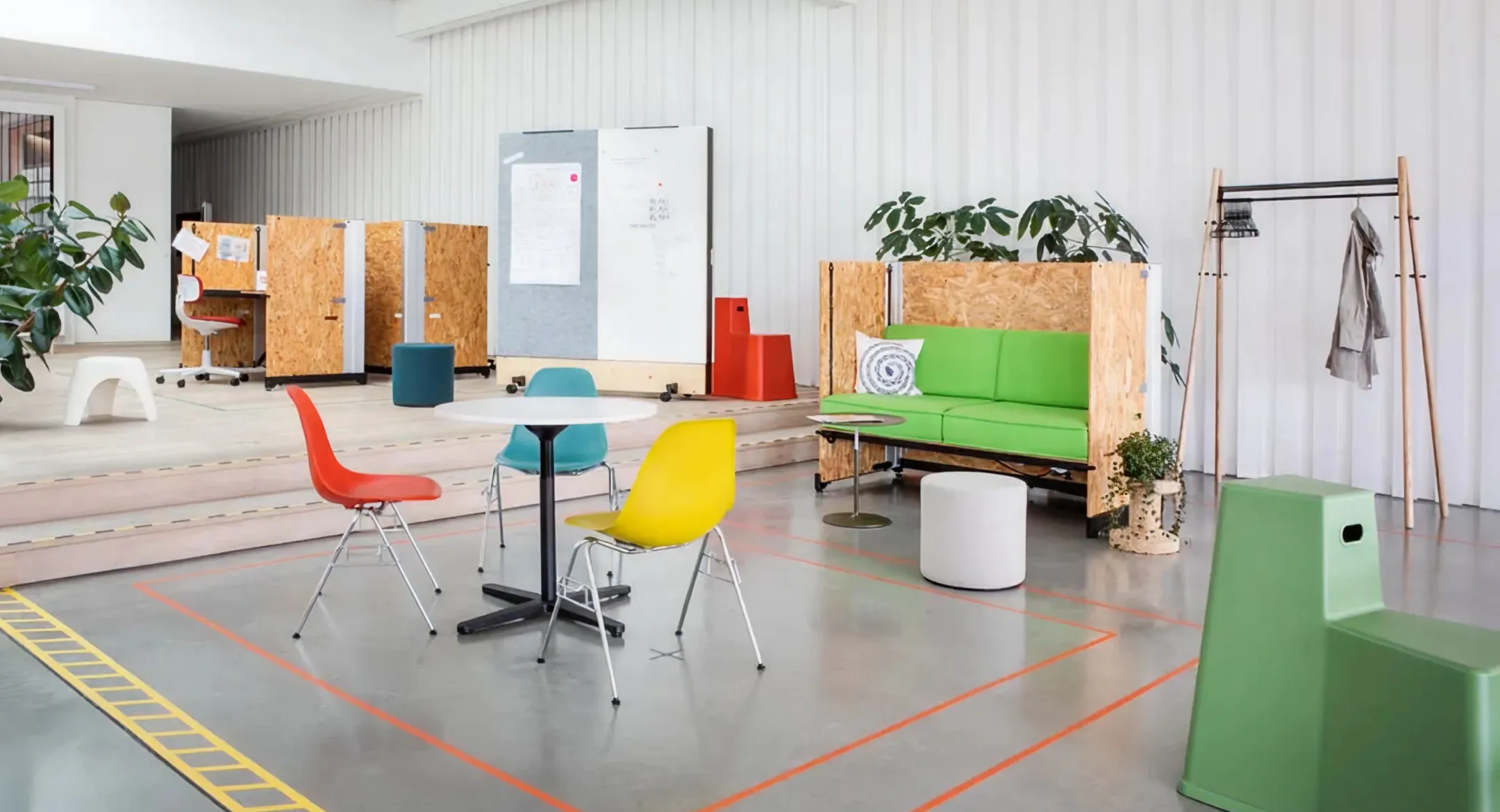 Modern office lounge area with colorful chairs, a round white table, green sofa with wooden frame, plants, and coat rack.