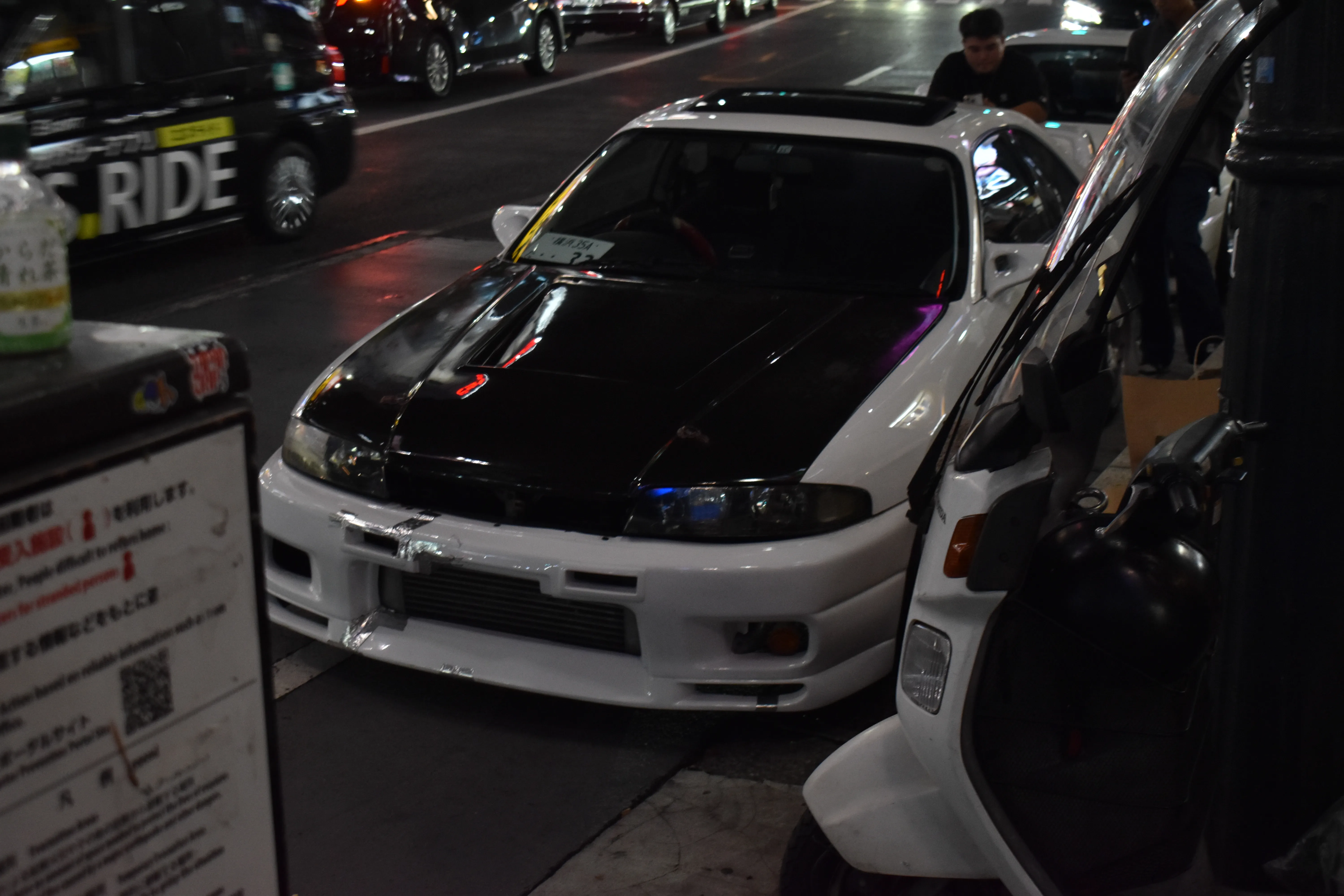 White and black Nissan Skyline car parked on a city street at night with people and other vehicles nearby. Shibuya Tokyo, Japan, the Pineworks vibe.