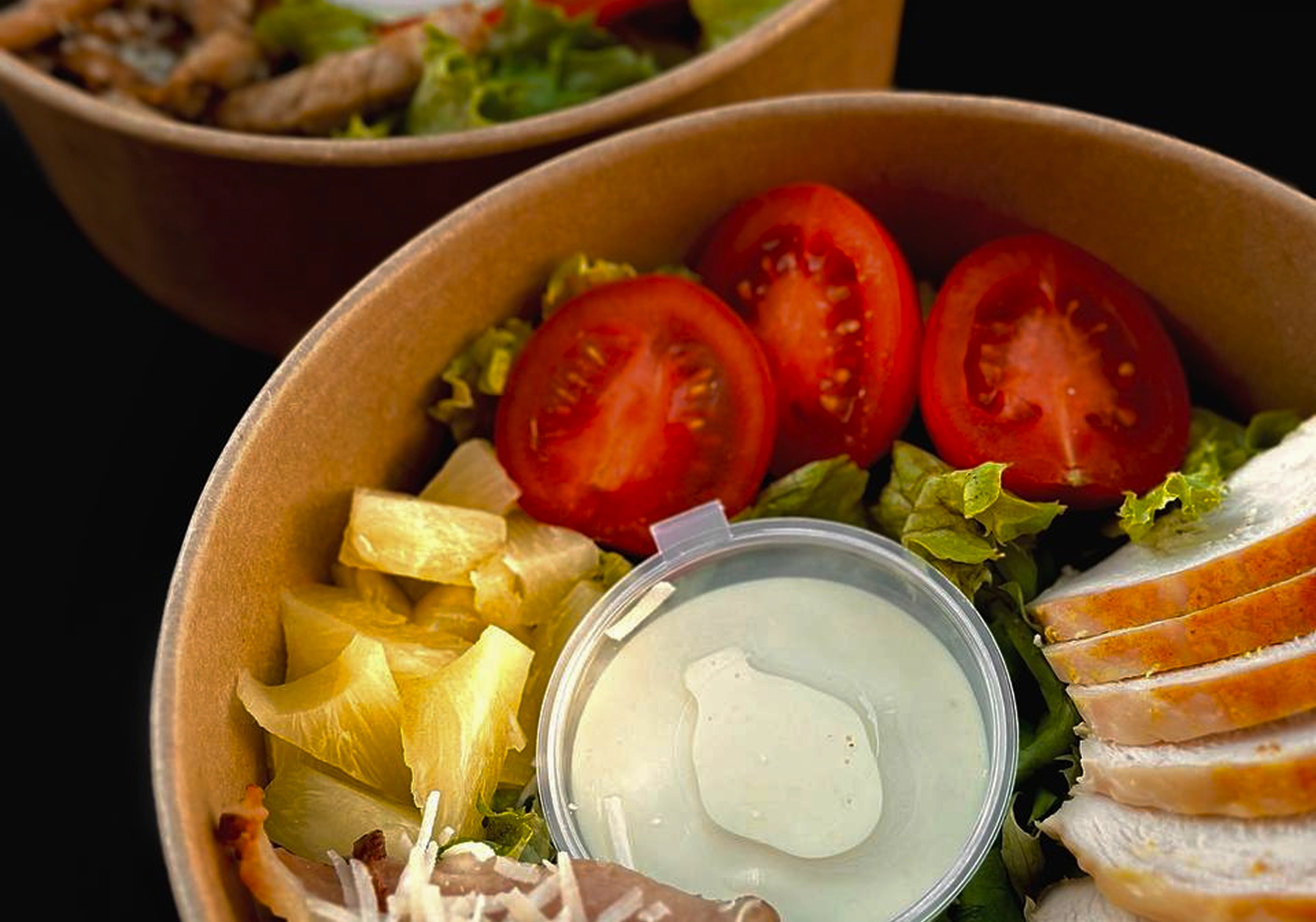 Close-up of a salad bowl with sliced tomatoes, pineapple chunks, grilled chicken slices, lettuce, and a container of creamy dressing.
