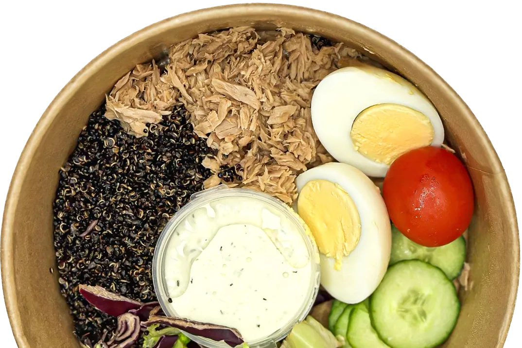 Bowl with black quinoa, shredded tuna, sliced cucumber, cherry tomato, halved hard-boiled eggs, mixed greens, and a container of creamy dressing.