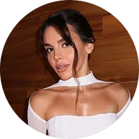 Young woman with dark hair styled in loose waves wearing a white off-shoulder top against a wooden background.