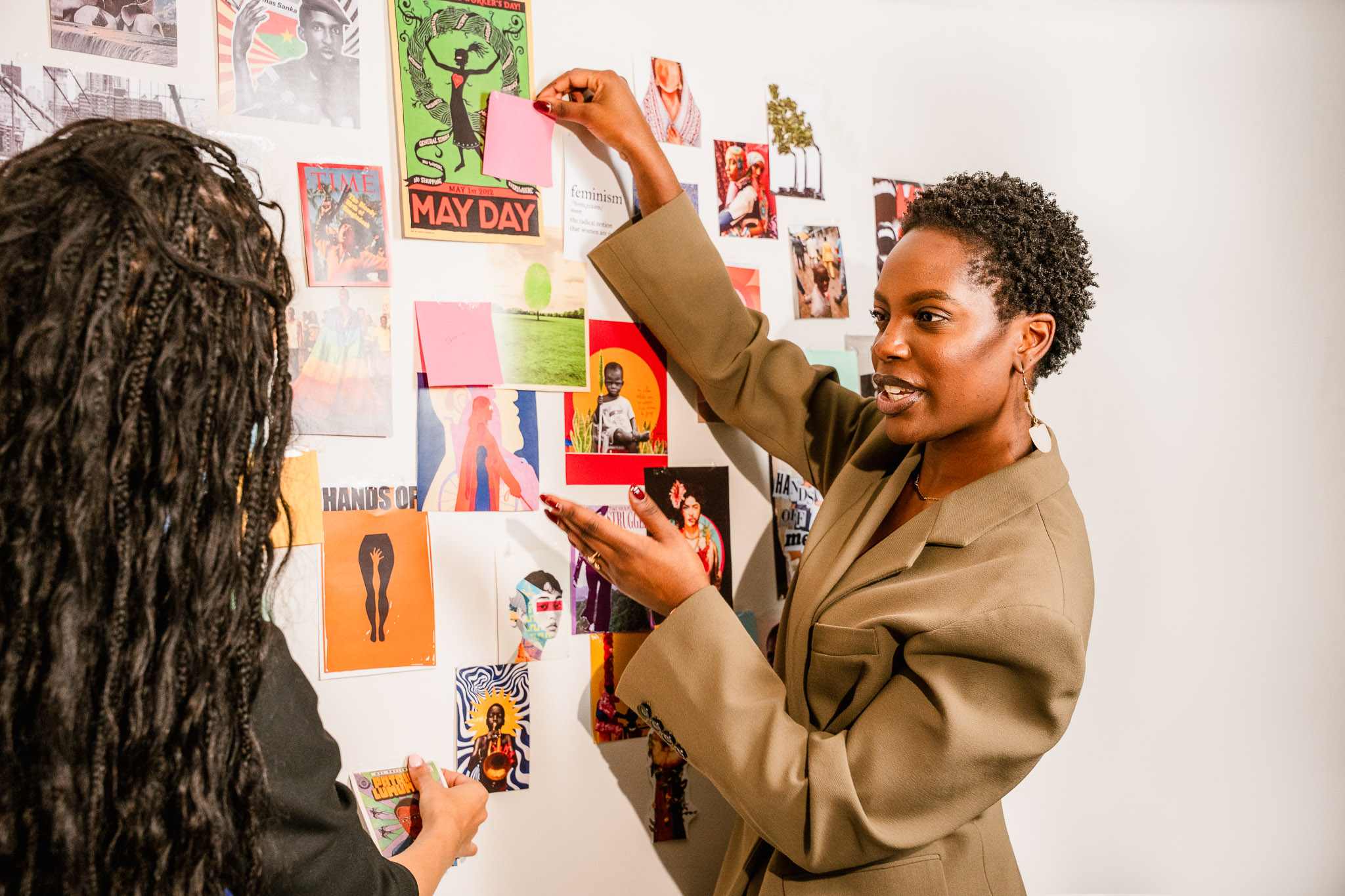 Two women discussing and arranging colorful posters and notes on a white wall.