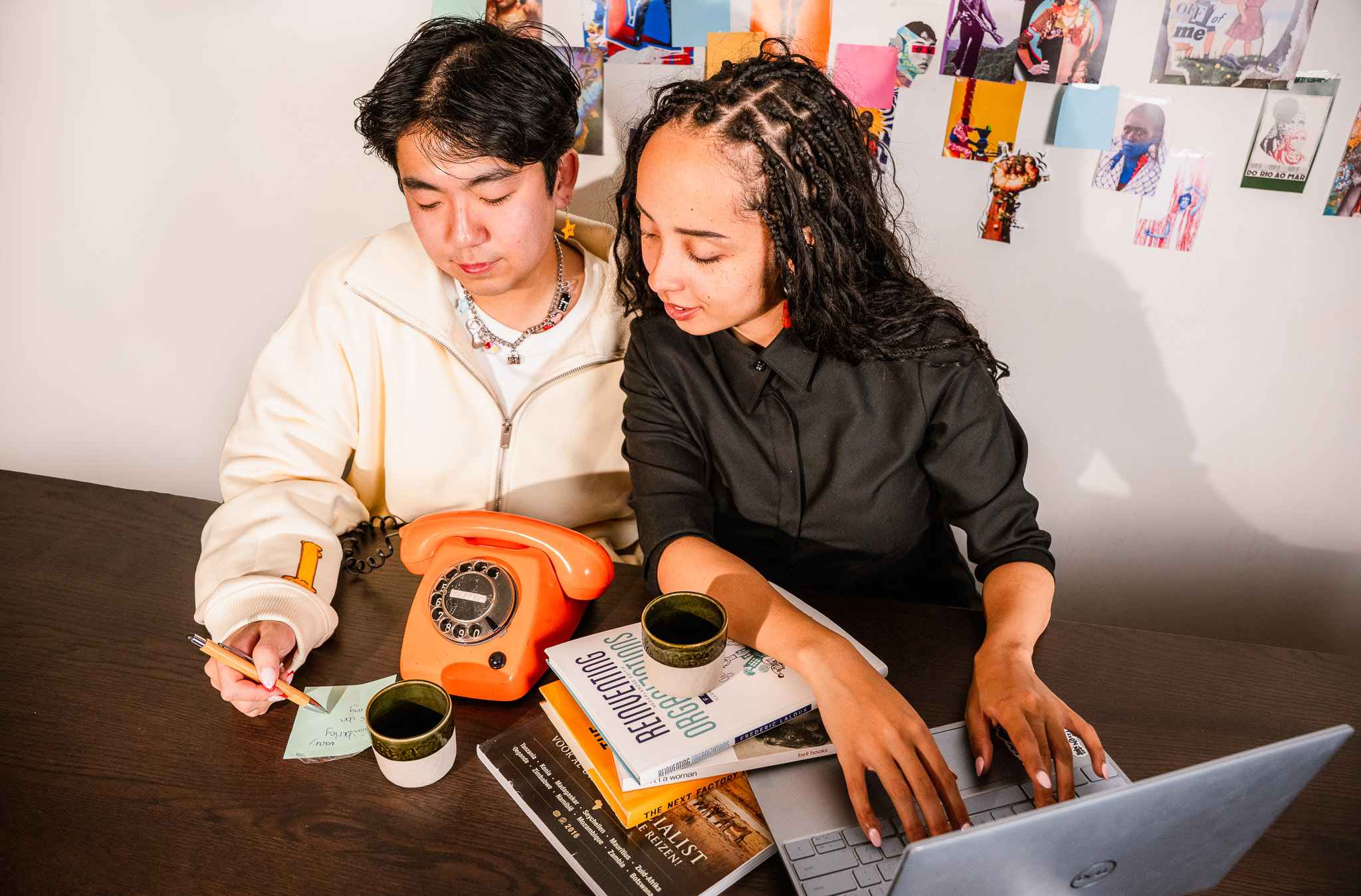Two people working together at a table with a laptop, books, notes, and an orange rotary phone.