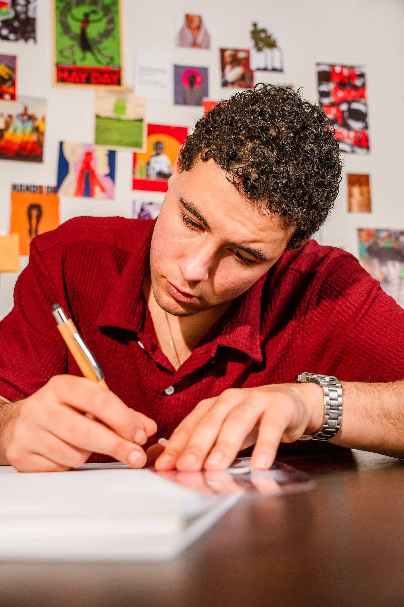 Young man in red shirt focusing intently while writing on paper at a desk with colorful posters on the wall behind.