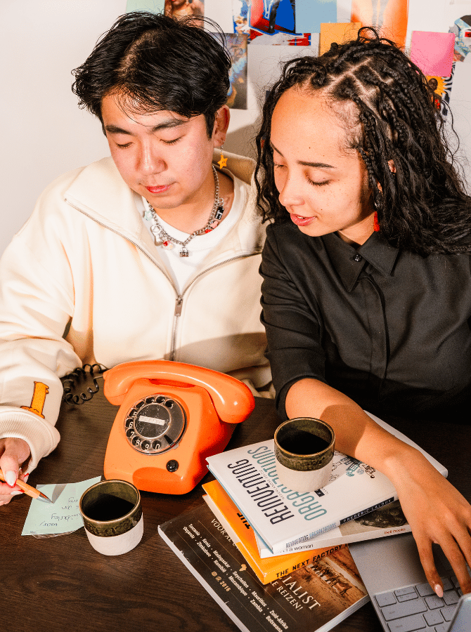 Two people working together at a table with a laptop, books, notes, and an orange rotary phone.