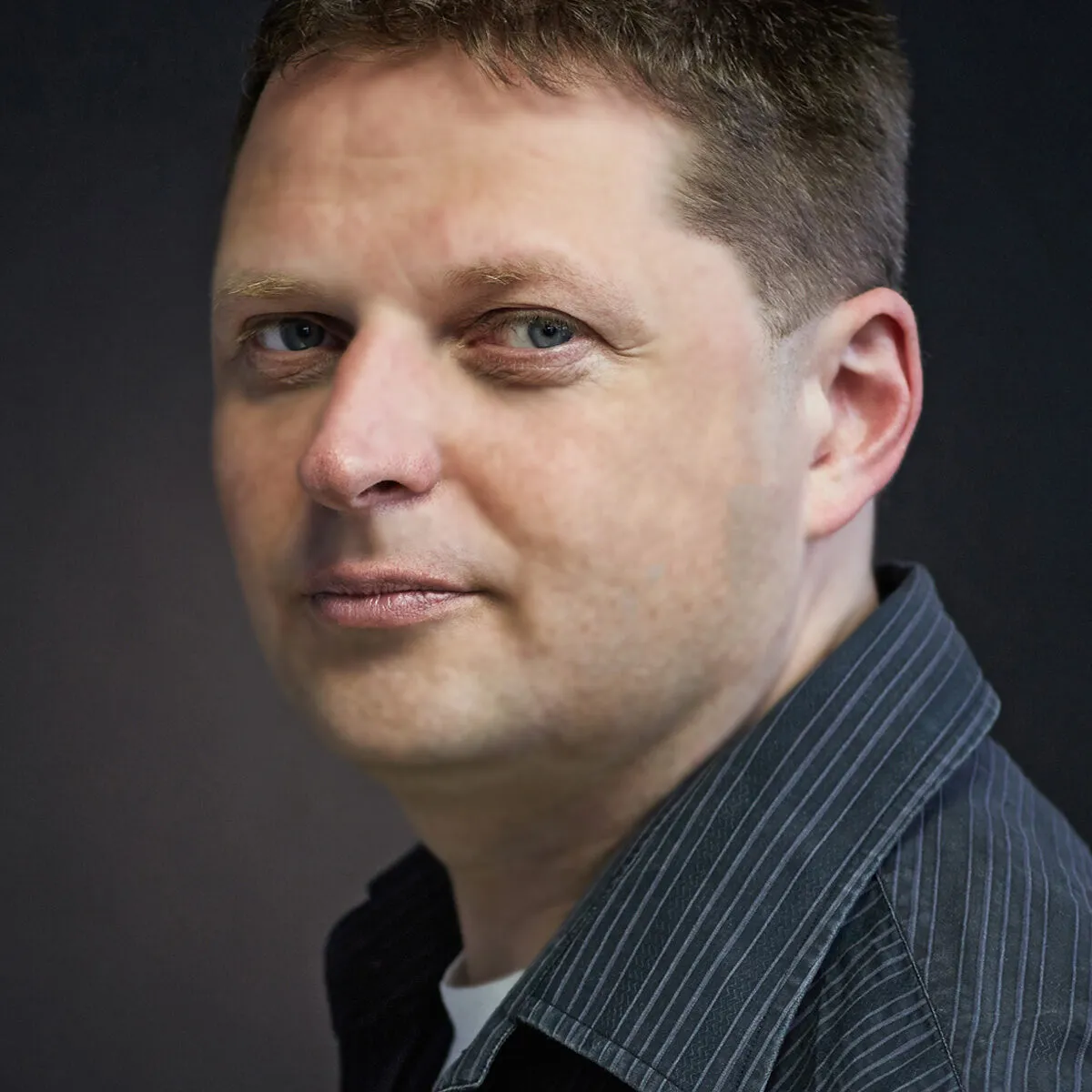 Close-up portrait of a man with light skin, short brown hair, and blue eyes against a dark background.