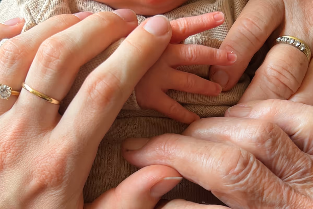 Close-up of three generations’ hands with wedding rings gently touching a baby's hand on a beige knitted outfit.