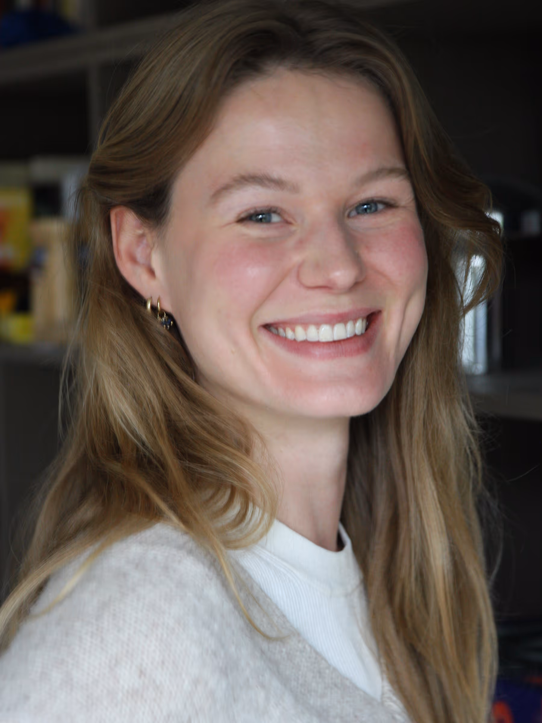 Smiling woman with long blonde hair wearing gold earrings and a beige sweater.