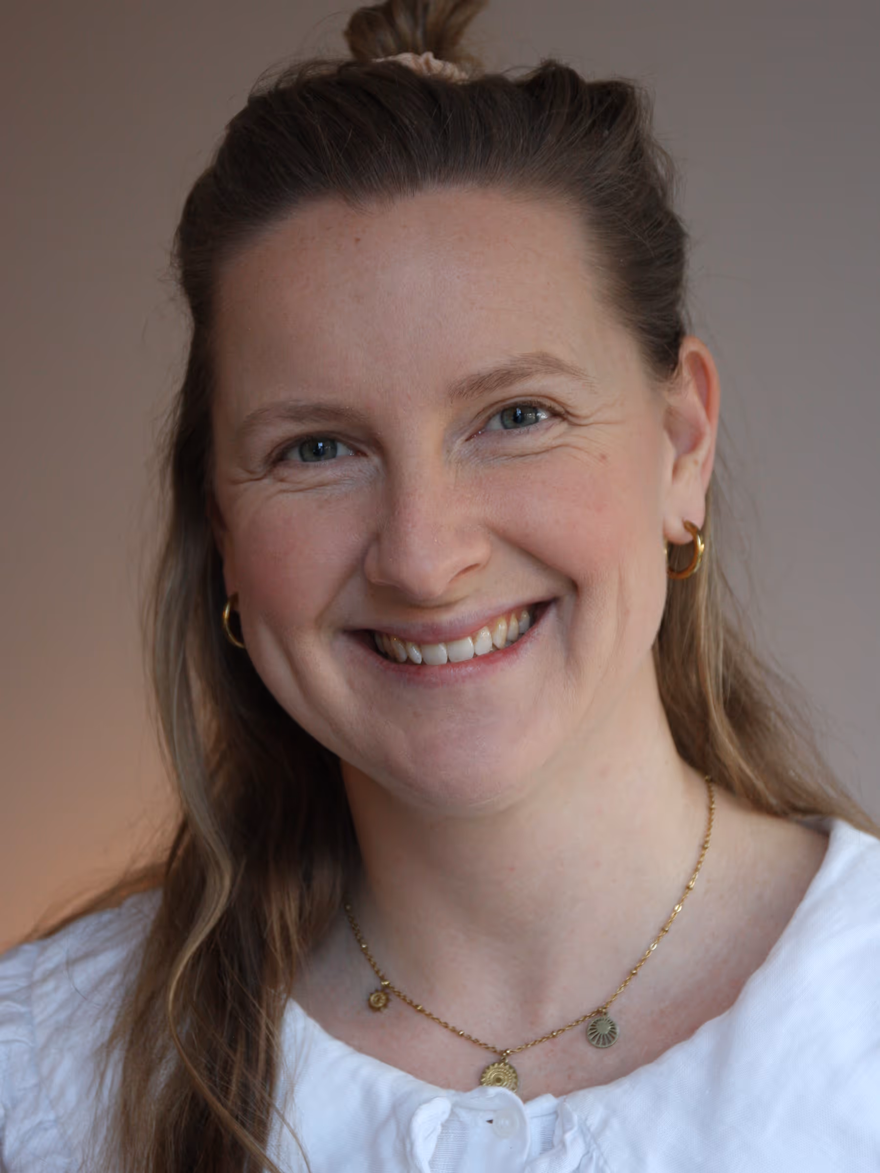 Smiling woman with light brown hair tied back, wearing gold hoop earrings, a gold necklace, and a white top.