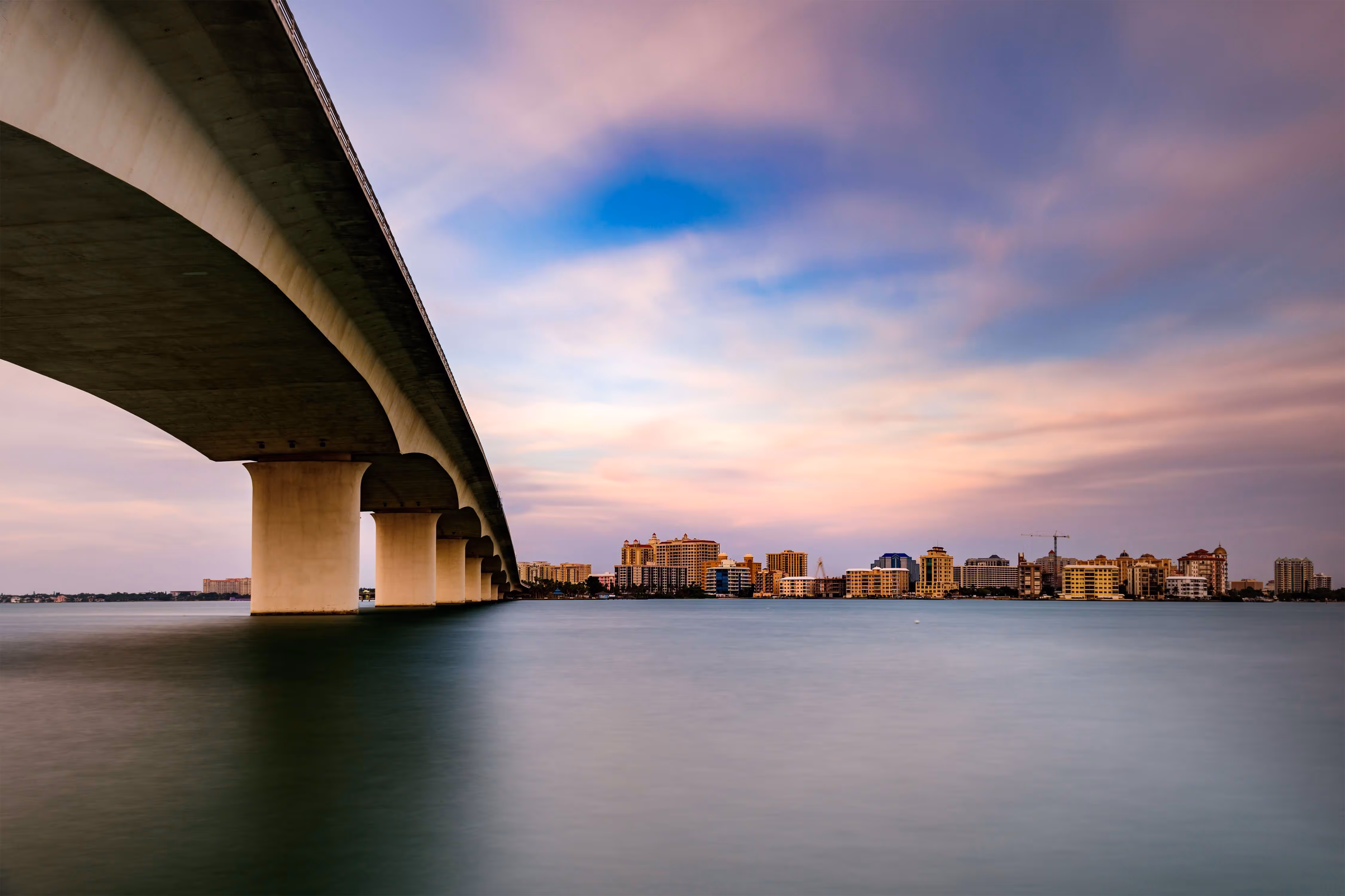 Wide concrete bridge extending over calm water toward a city skyline under a colorful sunset sky.