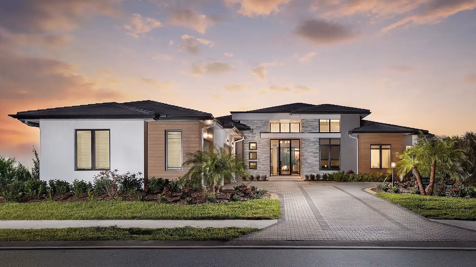 Modern single-story house with stone, wood, and white exterior at sunset with landscaped front yard and paved driveway.
