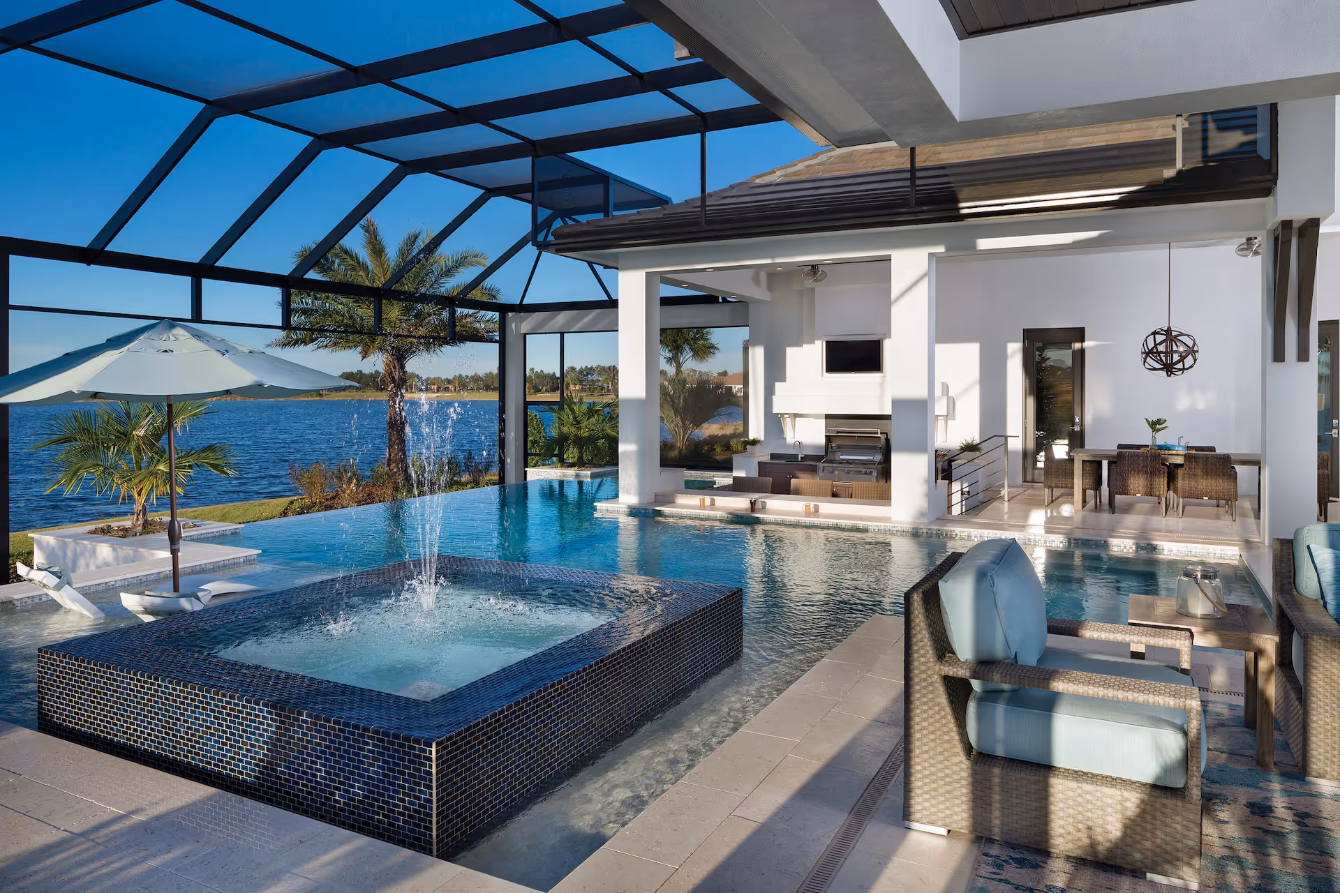 Modern outdoor living space with a screened pool, square hot tub with water jets, lounge chairs under umbrella, and a covered patio with dining and seating areas overlooking a lake.