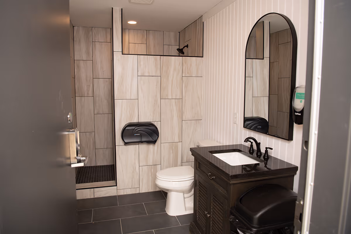 Modern bathroom with black countertop vanity, round mirror, white toilet, tiled shower, and soap dispenser on wall.