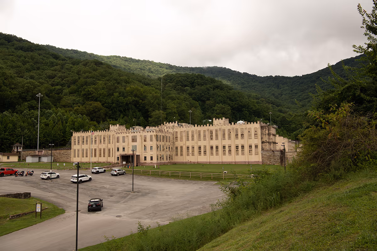 Large beige prison building surrounded by greenery and mountains under cloudy sky.