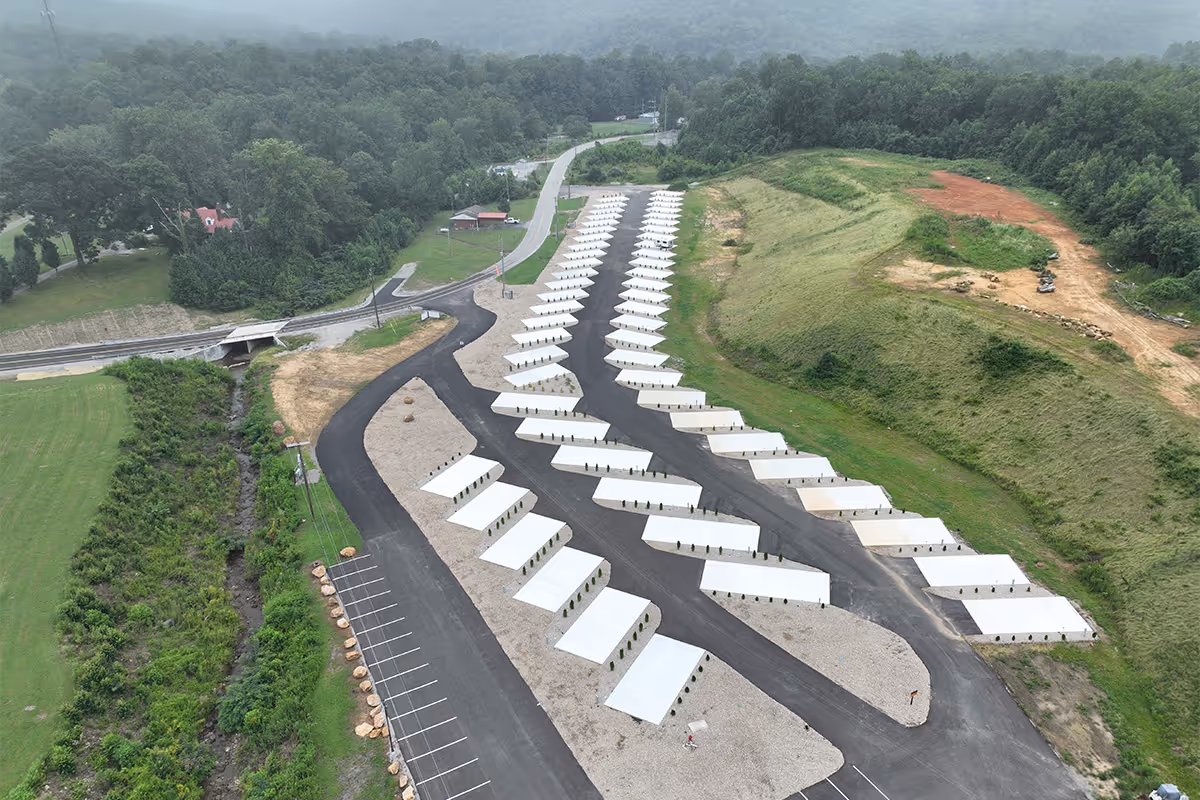 Aerial view of a campground with rows of paved and gravel areas, surrounded by green grassy hills and forest.