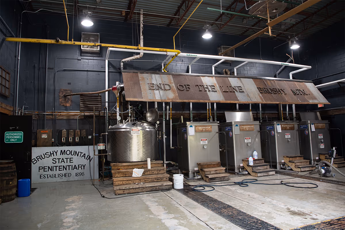 Industrial interior with metal fermenting tanks and equipment under a large rusted sign reading 'END OF THE LINE BRUSHY MTN', and a wall sign that says 'Brushy Mountain State Penitentiary Established 1896'.