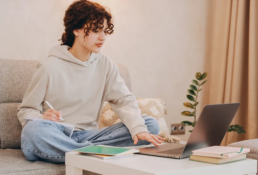 Young woman sitting cross-legged on a couch, writing in a notebook while using a laptop on a coffee table.