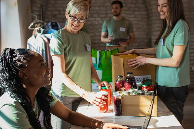 Three volunteers sorting jars of food and packing donation boxes in a community center.