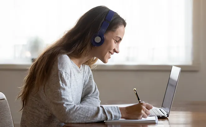 Teen female wearing headphones smiling while writing notes beside a laptop.