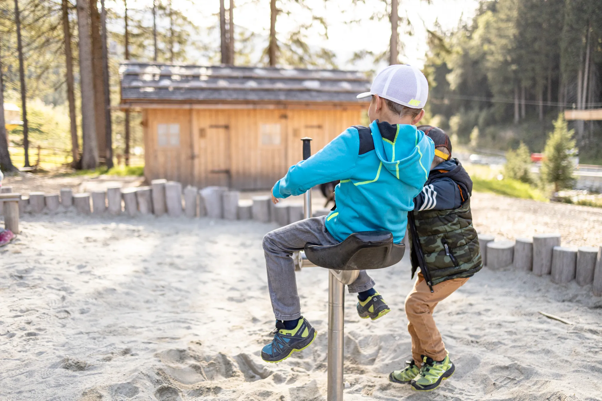 Zwei Jungen spielen auf einem Karussell im Sand vor einer Holzhütte im Wald.