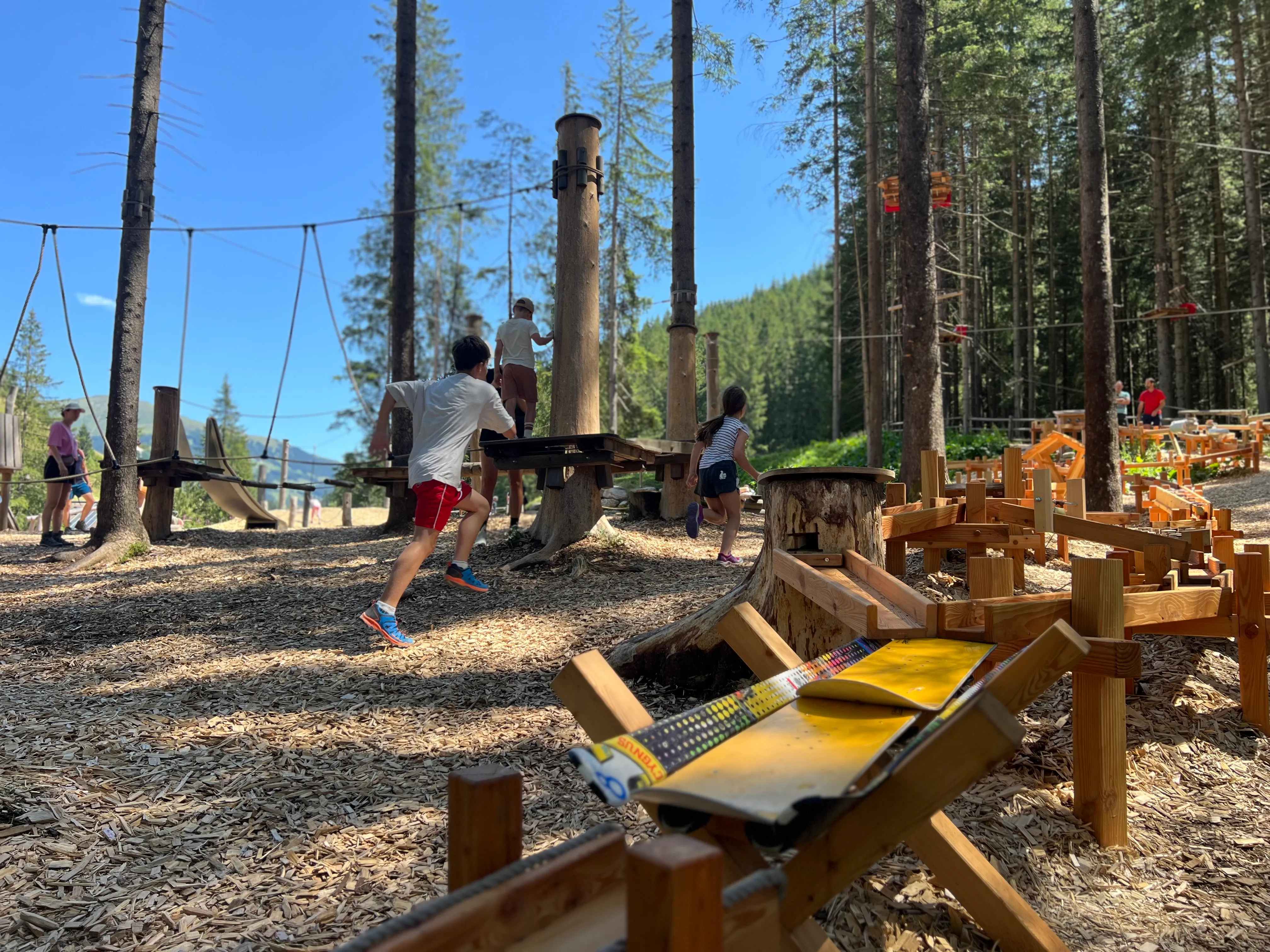 Kinder spielen im Wald auf einem Abenteuer-Spielplatz mit Holzstrukturen und Seilrutschen unter blauem Himmel.
