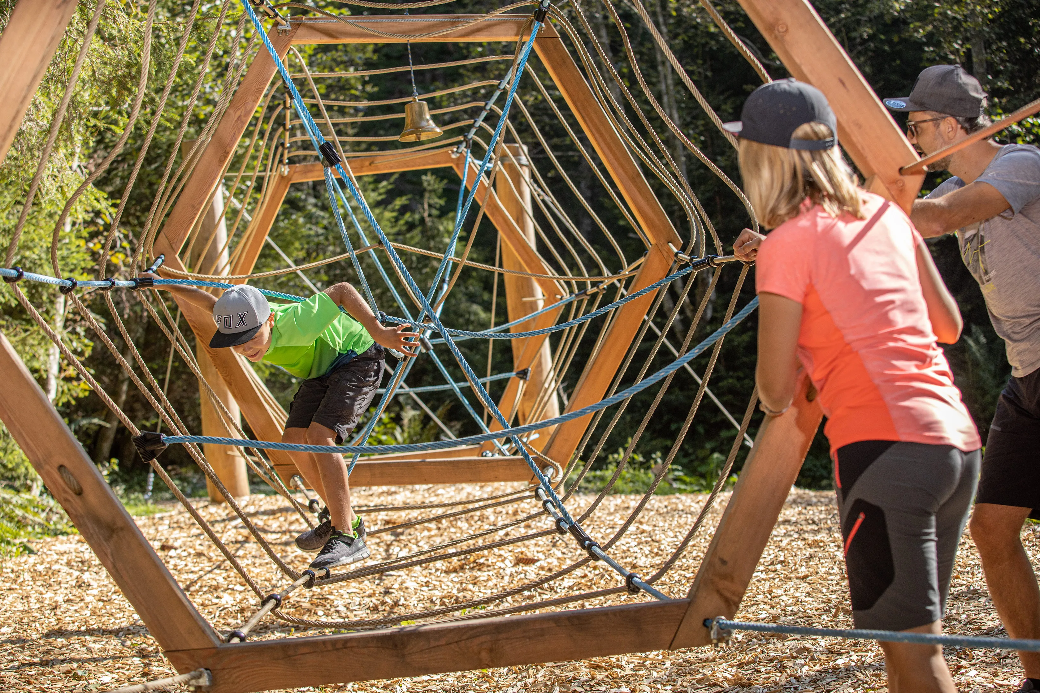 Junge klettert im Waldspielplatz durch ein seilgewebtes Klettergerüst aus Holz, zwei Erwachsene beobachten ihn.