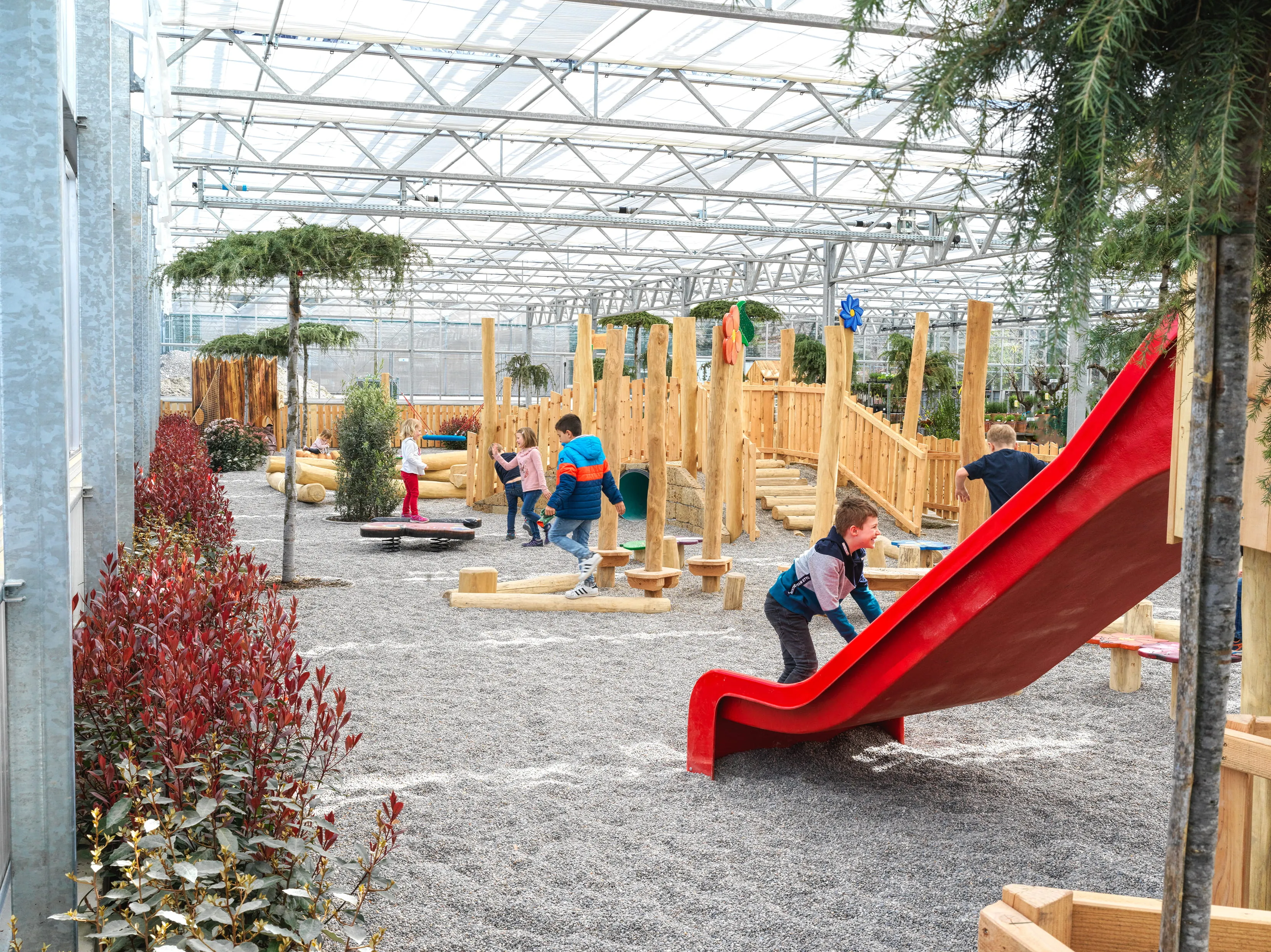 Innenbereich eines Kinderspielplatzes mit mehreren spielenden Kindern, Holzklettergerüsten und einer roten Rutsche unter einem Glasdach.