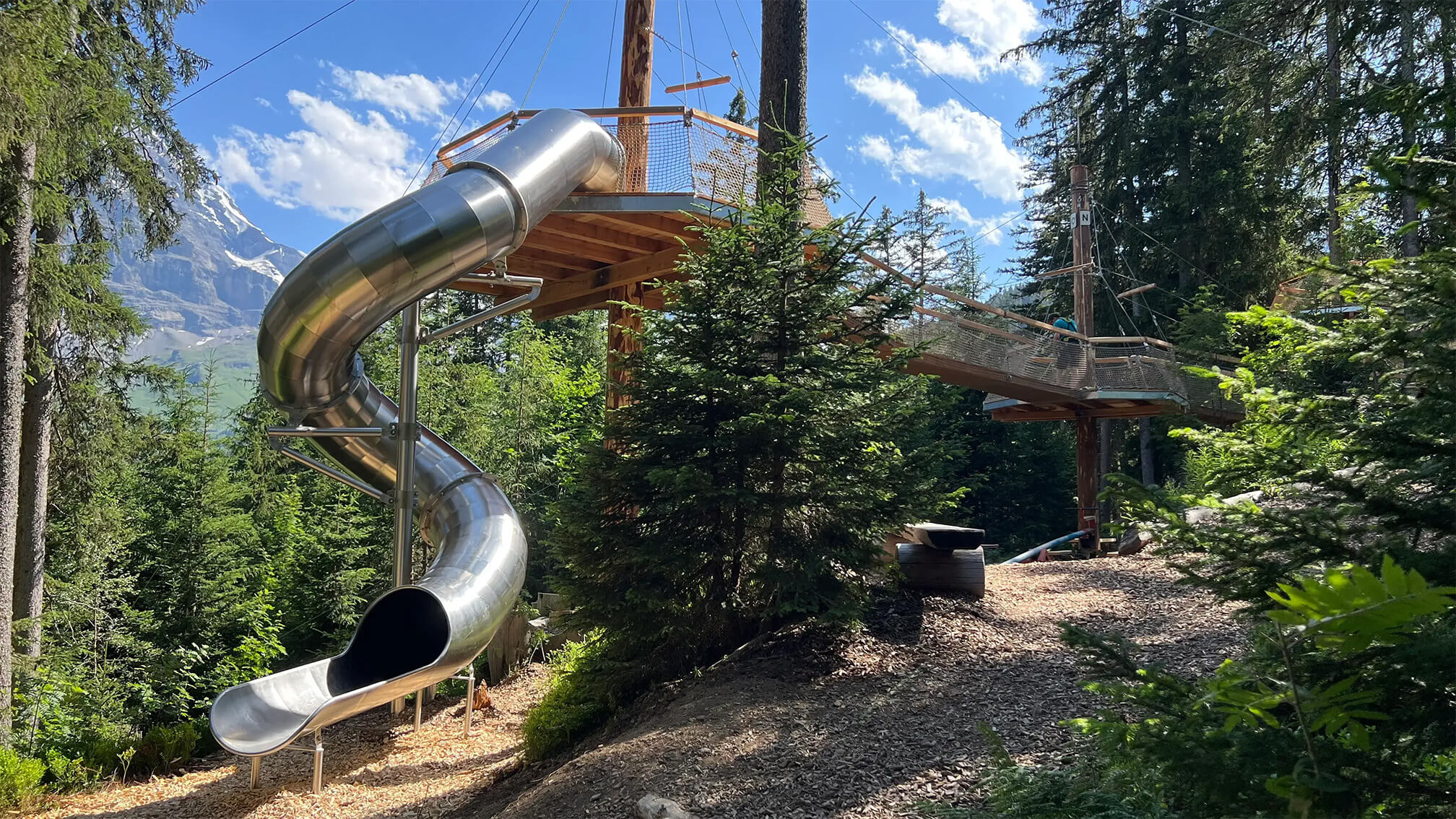 Glatte, spiralförmige Edelstahlrutsche mit Aussichtsplattform im Wald vor Bergkulisse.