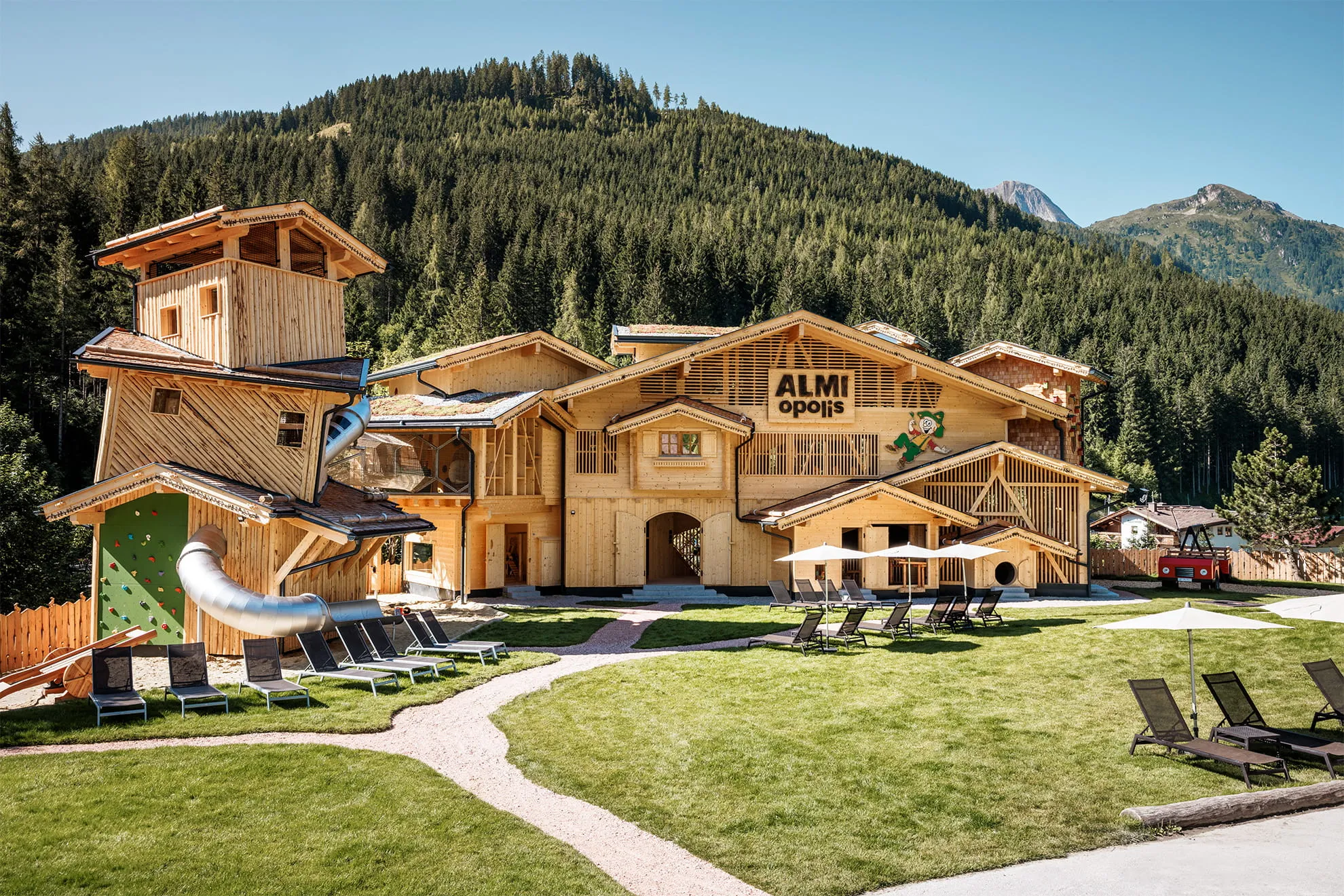 Holzgebäude ALMI opolis mit Rutsche, Kletterwand und Liegestühlen vor einem bewaldeten Berg im Zillertal.