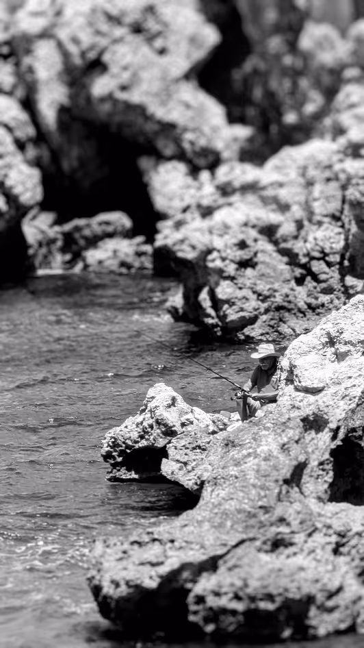 Person wearing a wide-brim hat fishing while sitting on rocky shoreline by calm water.