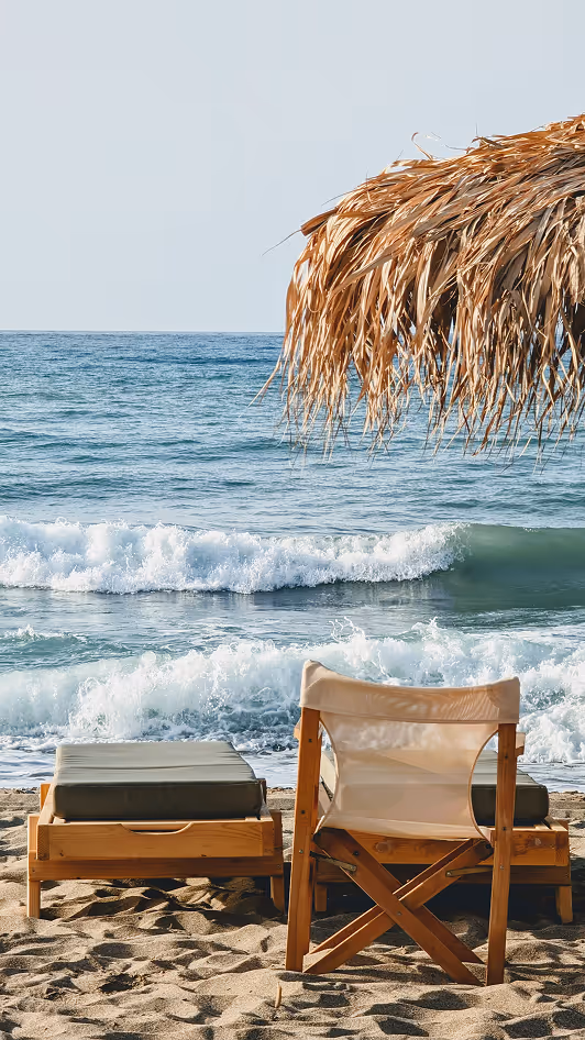Beach scene with two wooden lounge chairs and a thatched umbrella facing the ocean waves.