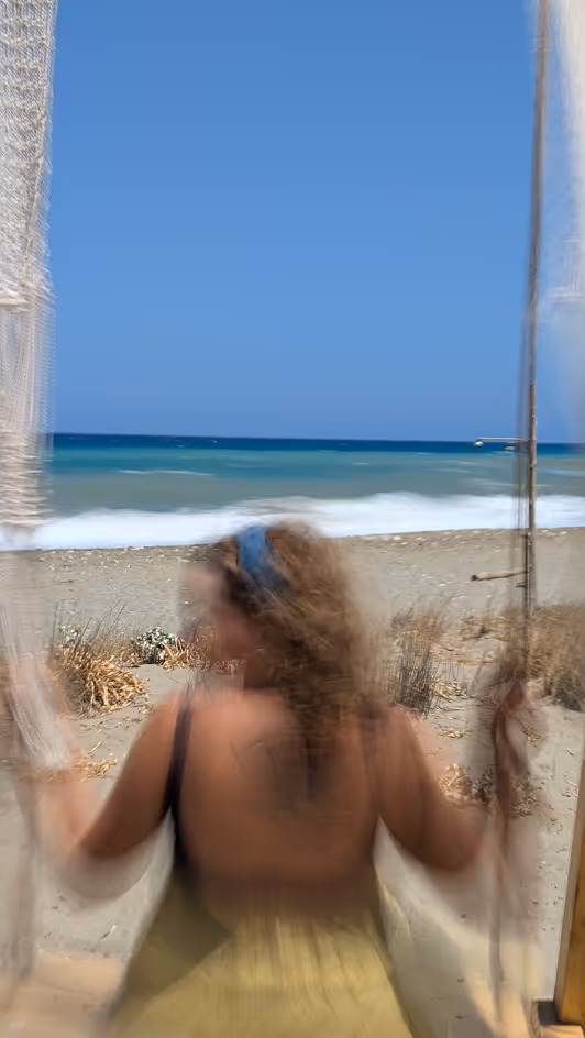 Blurred woman with a blue headband swings facing the ocean on a sandy beach under a clear sky.