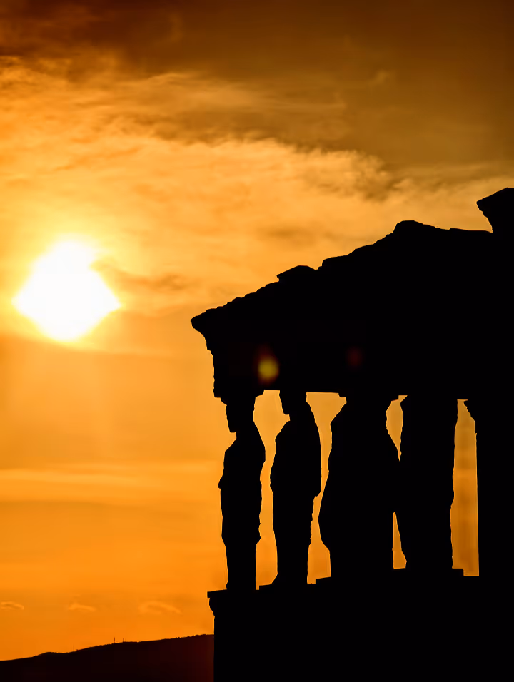 Silhouette of ancient Caryatids columns against an orange sunset sky with the sun visible.