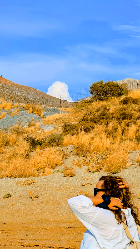 Person with curly hair and sunglasses in a white shirt in dry grassy landscape under blue sky.