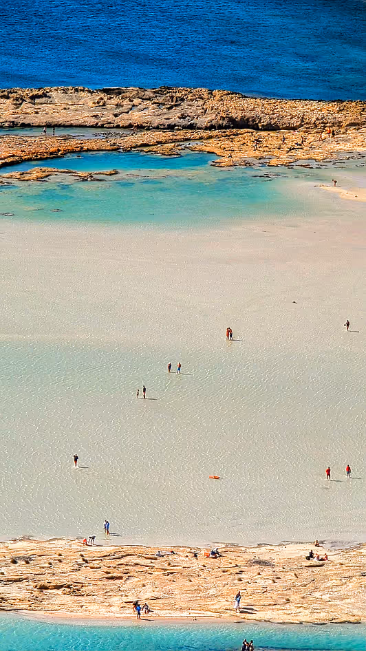People walking and relaxing on a shallow sandy beach with rocky edges and blue ocean water.