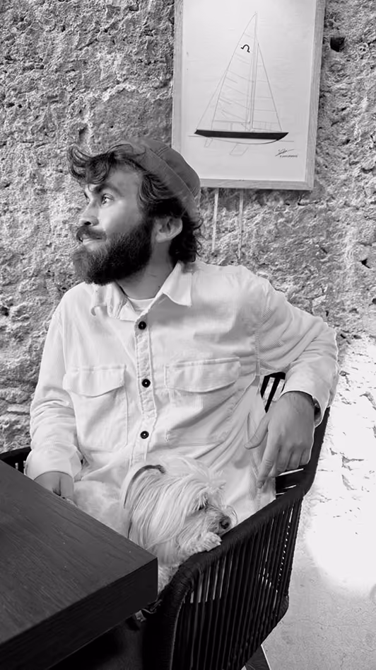 Bearded man wearing a beanie and button-up shirt sits in a chair beside a table with a small dog resting on his lap, against a textured wal.