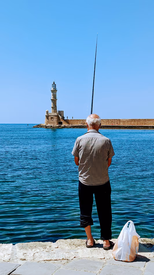 An elderly man with short gray hair fishing by the seaside, standing on stone pavement facing the water, with a lighthouse on a stone pier in the distance under a clear blue sky.