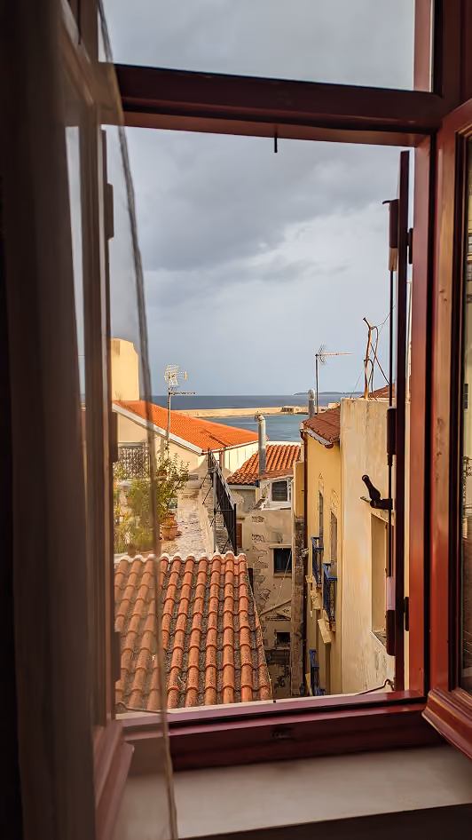 View through an open window with a sheer curtain, showing rooftops with red tiles, buildings with satellite antennas, and the sea under a cloudy sky in the distance.
