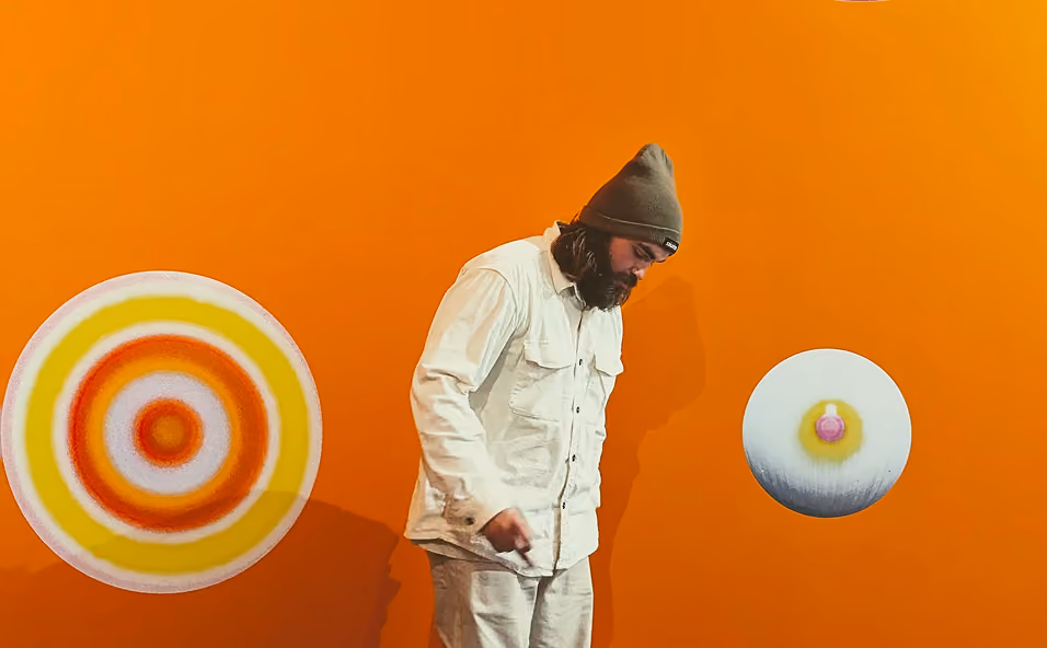 Man in white jacket and beige pants standing on a gray floor with an orange wall and circular art pieces.