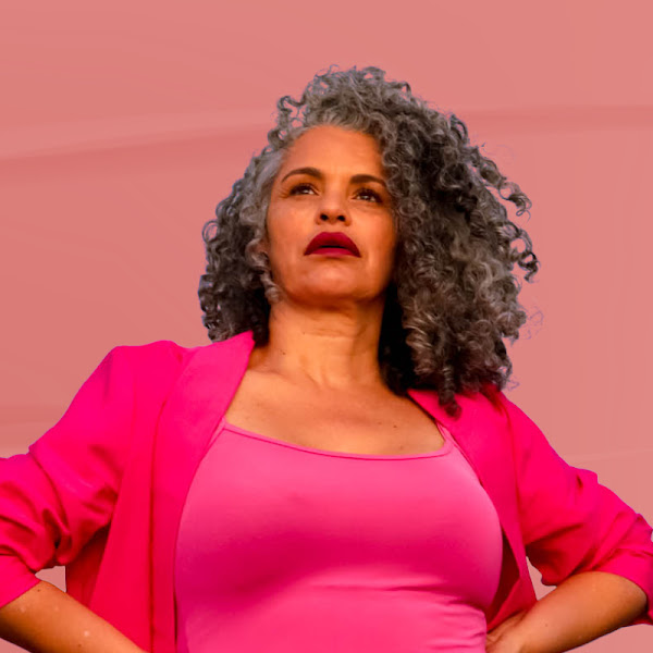 Confident woman with curly gray hair wearing bold pink top and jacket against a pink background.