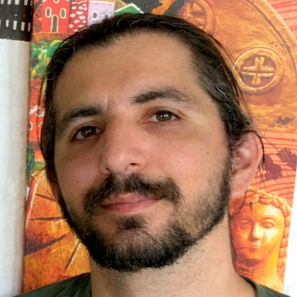 Close-up of a man with dark hair pulled back, a beard, and brown eyes, with colorful artwork in the background.