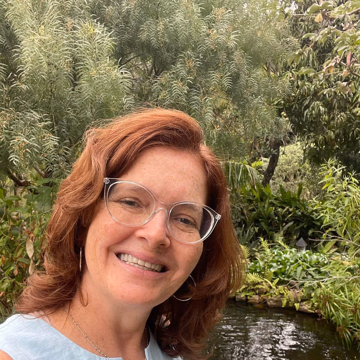 Smiling woman with red hair, clear glasses, and hoop earrings standing outdoors near a pond surrounded by greenery.