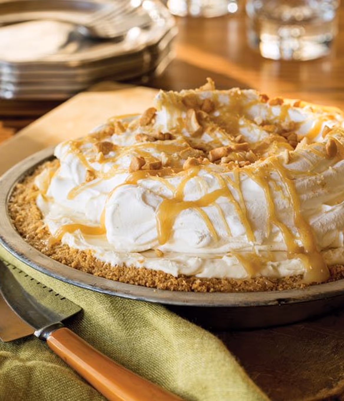 Creamy pie with a graham cracker crust, topped with whipped cream, caramel drizzle, and chopped nuts on a wooden table.