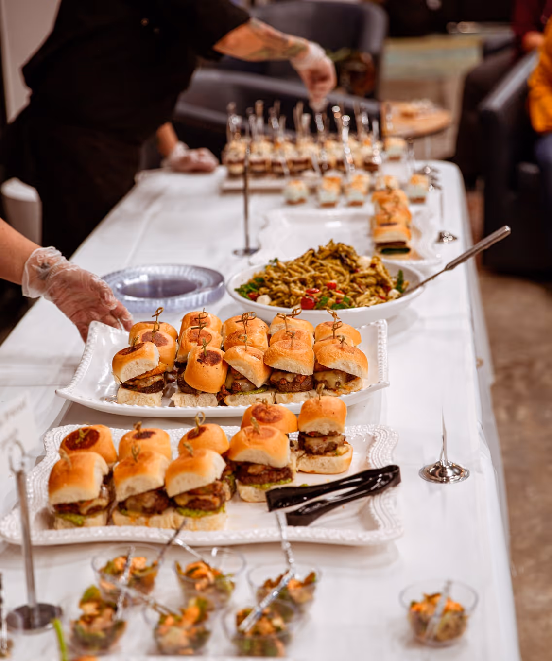 Table with trays of mini burgers and pasta salad being arranged by people wearing gloves at a catering event.