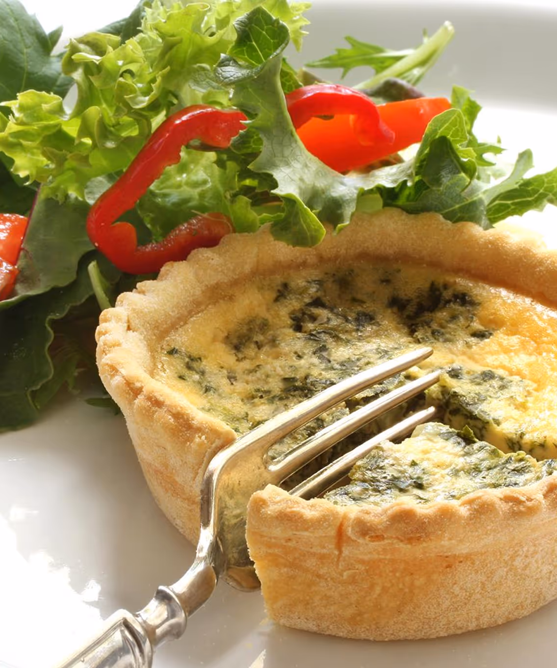 Spinach quiche slice on a white plate with fresh green leafy salad and red pepper strips, with a silver fork inserted into the quiche.